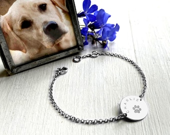 May include: A silver chain bracelet with a round charm that says "Charlie" and has a paw print. The bracelet is on a white surface with a framed photo of a dog in the background.