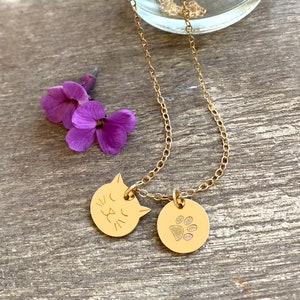 May include: A gold chain necklace with two charms. One charm is a round disc with a cat face engraved on it. The other charm is a round disc with a paw print engraved on it.