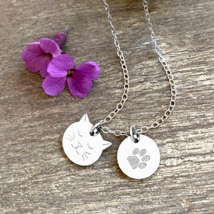 May include: A silver necklace with two charms, one shaped like a cat's face and the other a paw print. The necklace is on a wooden surface with purple flowers.