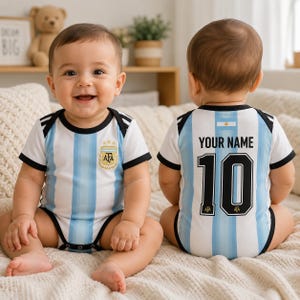Puede incluir: Un body para bebé con las rayas verticales azul claro y blancas de la camiseta de la selección argentina. El frente muestra el escudo de la AFA, mientras que la parte posterior presenta el texto YOUR NAME sobre el número 10 en negro y blanco.