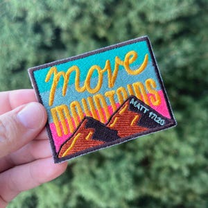 May include: A colorful embroidered patch with the text "Move Mountains" in yellow lettering and a mountain range graphic. The text "Matt 17:20" is also included in smaller lettering.