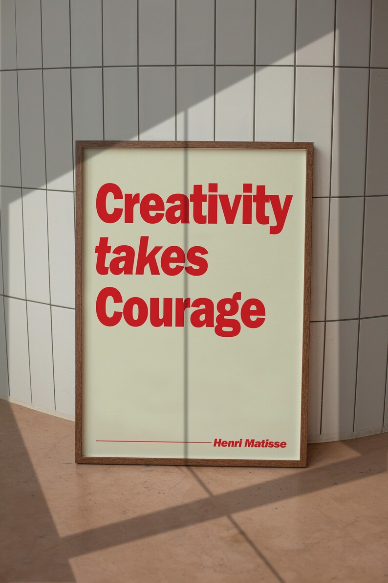 May include: A framed wall art print featuring a cream-colored background with bold red text that reads Creativity takes Courage and includes the name Henri Matisse at the bottom right, enclosed in a simple brown wooden frame.