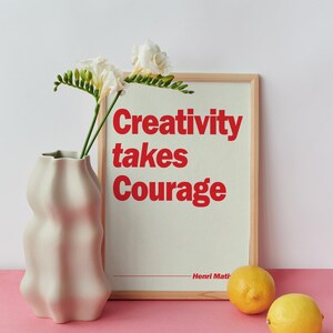 May include: A light wooden frame containing a print with the red text Creativity takes Courage and Henri Matisse, positioned next to a wavy-edged off-white ceramic vase holding white freesia flowers and two yellow lemons on a pink surface.