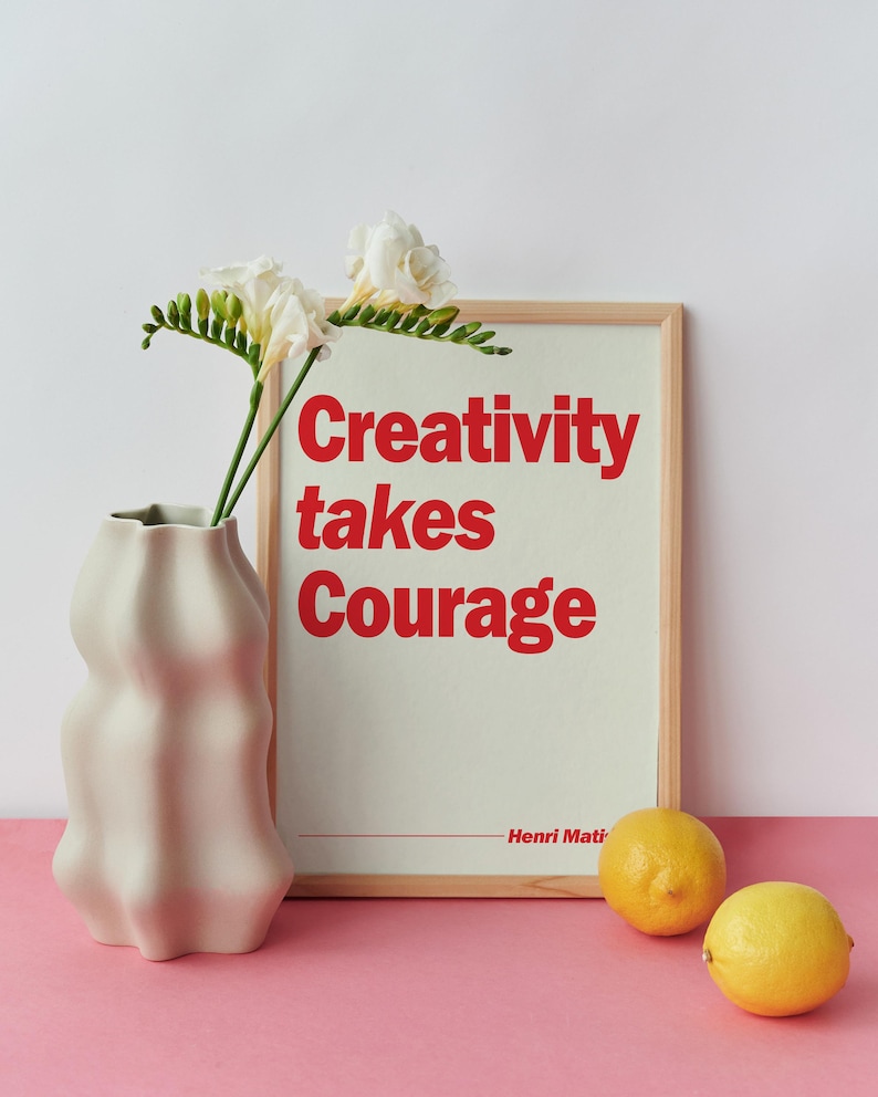 May include: A light wooden frame containing a print with the red text Creativity takes Courage and Henri Matisse, positioned next to a wavy-edged off-white ceramic vase holding white freesia flowers and two yellow lemons on a pink surface.
