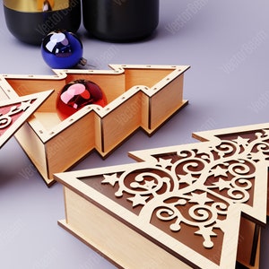 Peut inclure: Deux boîtes en bois en forme de sapin de Noël avec des découpes complexes. Une boîte est ouverte et contient une boule rouge. L'autre boîte est fermée et présente un design plus élaboré.