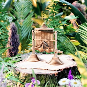 May include: A wooden sculpture of a complex, intricate design, resembling a miniature building or tower. The sculpture is made of many small, interlocking pieces of wood and is painted in a natural wood colour. It is sitting on a tree stump in a garden setting.
