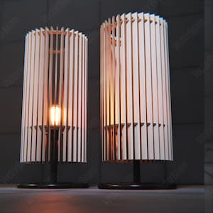 Könnte beinhalten: Zwei Holz-Tischlampen mit einem Lamellendesign und warmen Glühbirnen. Die Lampen haben schwarze Sockel und stehen auf einer grauen Oberfläche.