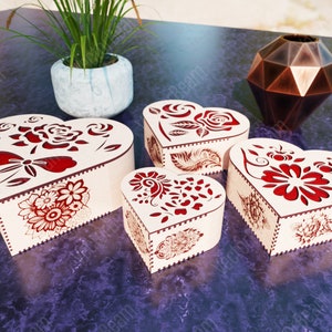 Pode incluir: Quatro caixas de madeira em forma de cora&ccedil;&atilde;o com intrincados desenhos florais vermelhos. As caixas est&atilde;o dispostas sobre uma superf&iacute;cie roxa.