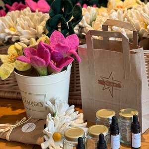 Pode incluir: Um conjunto de seis pequenos frascos de vidro com tampas douradas e conta-gotas, etiquetados com diferentes misturas de óleos essenciais. Os frascos estão dispostos sobre uma superfície de madeira com um saco de papel castanho, um ramo de flores brancas e um balde de lata branco com a etiqueta "Flowers".