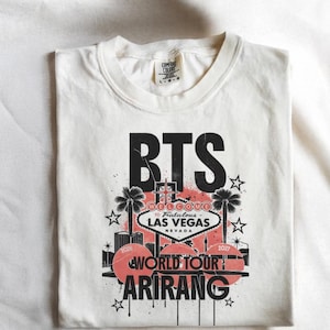 Puede incluir: Camiseta color crema con un diseño gráfico. El diseño incluye el texto "BTS" en negro, un letrero de Las Vegas, palmeras, estrellas y las palabras "World Tour Arirang".