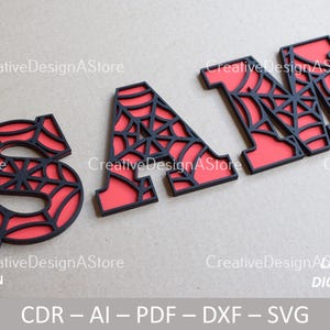 Könnte beinhalten: Ein Satz aus drei schwarzen und roten Buchstaben, die "SAM" buchstabieren, mit einem Spinnennetz-Design. Die Buchstaben sind aus Holz ausgeschnitten und haben einen roten Hintergrund.