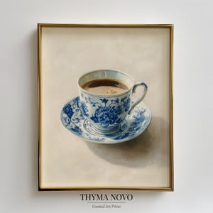 May include: A framed print of a teacup and saucer filled with coffee. The teacup and saucer are white with blue floral patterns. The print is in a gold frame and has the text "THYMA NOVO Curated Art Prints" at the bottom.