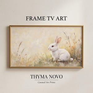 Op de afbeelding: Een ingelijste kunstdruk met een wit konijn in een veld met hoog gras en wilde bloemen. Het kunstwerk heeft een zacht, pastelkleurig kleurenpalet met de tekst "FRAME TV ART" en "THYMA NOVO" bovenaan en onderaan.