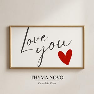 May include: A framed art print with the words "Love you" in black script, accompanied by a red heart. The print is set against a white background and framed in a gold-colored frame. The bottom of the print includes the text "THYMA NOVO" and "Curated Art Prints".