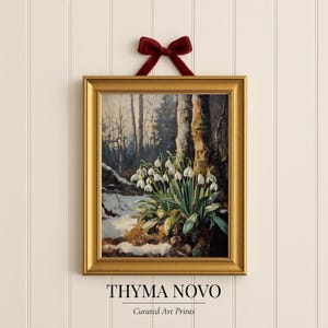 以下が含まれることがあります： 雪解けの森の風景を描いた額入りのアートプリント。前景にはスノードロップが描かれています。絵画は金のフレームに入っており、赤いベルベットのリボンで吊り下げられています。「THYMA NOVO Curated Art Prints」の文字が下部にあります。
