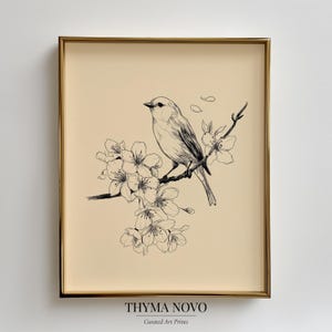 May include: A framed art print featuring a detailed black and white illustration of a bird perched on a flowering branch. The artwork is set against a beige background and framed in a gold-colored frame. The text "THYMA NOVO Curated Art Prints" is at the bottom.
