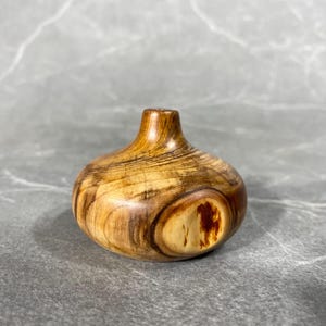 Puede incluir: Un pequeño jarrón de madera con forma redonda y bulbosa. El jarrón está hecho de madera marrón claro con vetas y nudos marrones más oscuros. Tiene un acabado liso y pulido.