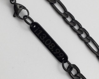 May include: Black Figaro chain necklace with a rectangular pendant engraved with the date "13.08.99". The necklace is made of a dark metal and features a lobster clasp closure. The chain has a classic, elongated oval link design.