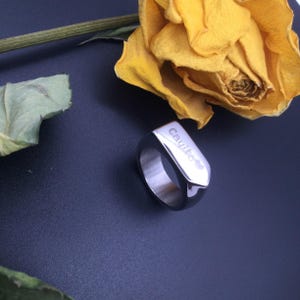 May include: A silver-colored ring with the name "Cayito" engraved on a rectangular surface, accompanied by a small heart symbol. The ring is set against a dark background with a yellow rose.