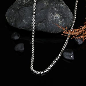 May include: A silver-toned box chain necklace featuring a series of interconnected square links that form a smooth, continuous strand. The metallic chain has a polished finish that reflects light along its length, creating a bright, uniform appearance.