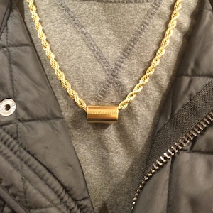 Könnte beinhalten: Eine goldfarbene Seilkette mit einem zylindrischen Anhänger. Der Anhänger hat eingravierten Text. Die Kette wird über einem grauen Hemd und einer schwarzen Steppjacke getragen. Schmuck.