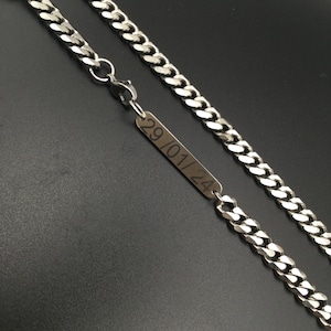 Puede incluir: Un collar de cadena de plata con una etiqueta rectangular. La etiqueta está grabada con la fecha "29/01/24". La cadena está hecha de eslabones entrelazados y tiene un cierre de mosquetón. El collar se muestra sobre un fondo oscuro.