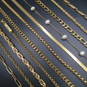 以下が含まれることがあります： 暗い背景に展示された、さまざまなゴールドカラーのネックレス。 ネックレスは、カーブチェーン、ロープチェーン、ヘリンボーンチェーンなど、さまざまなチェーンスタイルが特徴で、白い真珠で飾られたものもあります。 あるネックレスには「DEE」という文字が刻まれています。