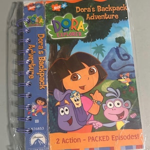 May include: A Dora the Explorer "Dora's Backpack Adventure" spiral-bound book. The cover features Dora, a purple backpack, and a monkey. The book is sealed in plastic and has the text "2 Action - PACKED Episodes!"