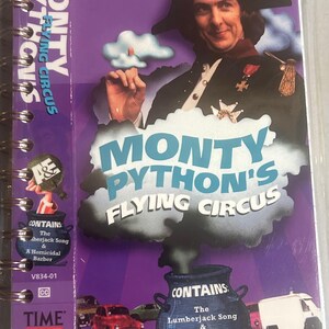 May include: A spiral-bound video case for Monty Python's Flying Circus with a purple background. The cover features a person in a historical military uniform, a blue cauldron with smoke, and text that reads: Contains: The Lumberjack Song & A Homicidal Barber.