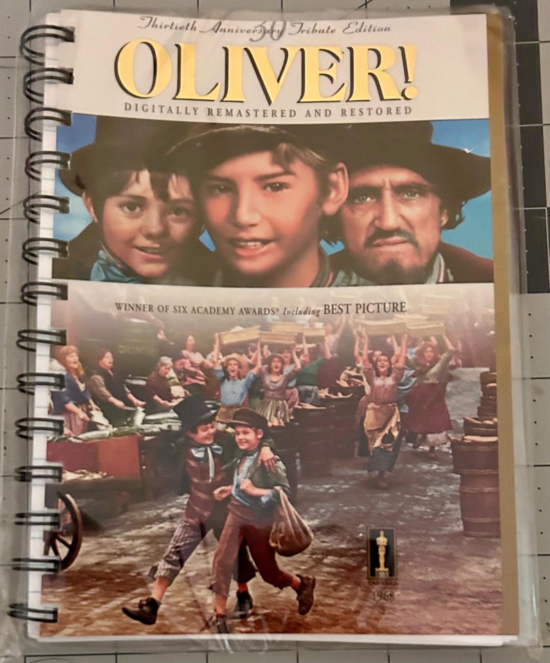 May include: A spiral-bound notebook featuring the movie poster for "Oliver!" The cover shows images of the cast and a scene from the film. Text includes "Oliver!" and "Digitally Remastered and Restored."