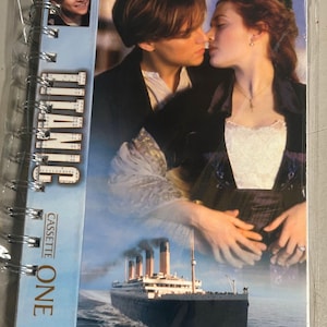 May include: A spiral-bound notebook featuring a Titanic movie cover with a romantic scene of two people, a small inset photo of a man, the text TITANIC, and the text CASSETTE ONE. Below the couple, the Titanic ship sails on the ocean.