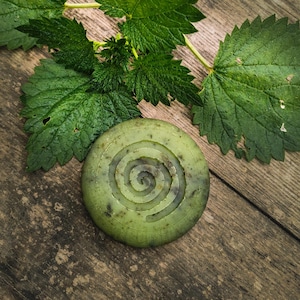 Puede incluir: Un jabón redondo verde con un diseño en espiral se encuentra sobre una superficie de madera envejecida. Hojas verdes frescas se disponen alrededor del jabón, creando una composición natural. El jabón es de un verde apagado y la madera tiene un aspecto rústico.