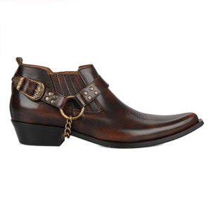 Puede incluir: Botas de tobillo de cuero marrón con punta puntiaguda y detalle de cadena. Las botas tienen una hebilla y una correa con tachuelas metálicas.