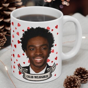 May include: White ceramic mug with a red heart pattern. The mug features a photo of a person with the name "Caleb McLaughlin" printed on a banner.