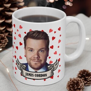 May include: White ceramic mug with a red heart pattern and a photo of James Corden. The mug has a black handle and the text "JAMES CORDEN" is printed on the bottom.