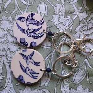 May include: Two circular keyrings with cream ceramic discs decorated with blue bird designs. Each keyring has a silver metal ring and clasp, and a small blue bead. The background features a green floral pattern.