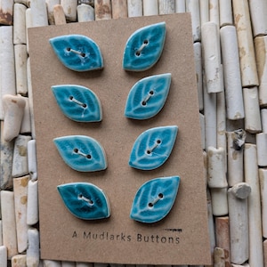 May include: Eight leaf-shaped ceramic buttons in shades of turquoise and teal with a glossy finish, arranged in two columns on a rectangular brown card. Each button features two holes and a debossed vein pattern. The text A MUDLARKS BUTTONS is printed.