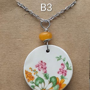 May include: A silver chain necklace with a circular, white pendant decorated with pink, orange, and green floral patterns. The pendant has an orange bead above it, and the text "B3" is visible.