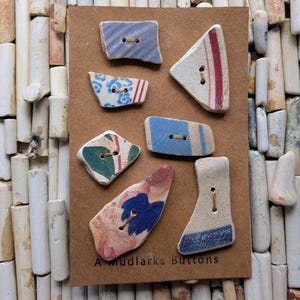May include: A collection of seven unique, irregularly shaped buttons with various patterns and colors, including blue stripes, floral designs, and red accents. The buttons are mounted on a brown card, with the text "A Mudlarks Buttons" at the bottom.