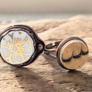 Könnte beinhalten: Zwei handgefertigte Ringe mit kupferfarbenen Metalleinfassungen. Ein Ring hat einen weißen Keramikmittelteil mit goldenem Blumendesign. Der andere Ring hat einen beigefarbenen Keramikmittelteil mit schwarzen geschwungenen Linien. Beide Ringe liegen auf einer Holzoberfläche.
