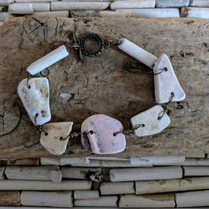 Pode incluir: Uma pulseira artesanal com fecho de alternância, composta por contas de cerâmica brancas, rosa e creme. As contas são conectadas com fio de cobre, criando uma joia única e natural. A pulseira é apresentada em uma superfície de madeira.