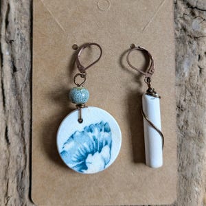 May include: Handmade earrings featuring a white ceramic disc with a blue floral design and a white cylindrical bead. The earrings have copper-colored ear wires and accents. One earring has a round ceramic bead, while the other has a white cylindrical bead.