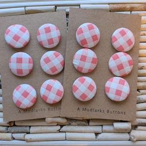 May include: Two sets of round fabric buttons with a pink and white gingham pattern. Each set of buttons is attached to a brown card with the text "A Mudlarks Buttons". The buttons are arranged in a grid pattern.