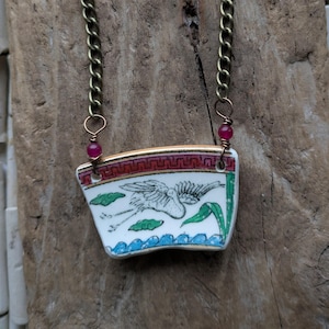 May include: A distinctive necklace with a porcelain pendant depicting a bird and floral pattern. The pendant, in white with red, green, and blue details, hangs from a gold-toned chain. Small, round, red beads are attached to the chain.