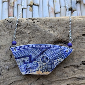 May include: A necklace featuring a fan-shaped pendant made from a fragment of blue and white patterned ceramic. The pendant is suspended from a silver-toned metal chain, accented with two small, round, solid purple beads on either side.