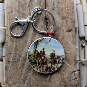 Puede incluir: Un llavero circular con una escena impresa de jinetes a caballo con chaquetas rojas y perros en un campo. El colgante está unido a una anilla de metal plateado y un cierre de mosquetón, acentuado por una pequeña cuenta roja y redonda.