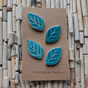 May include: Four turquoise leaf-shaped ceramic buttons with white veining, displayed on a brown card. The card reads "A Mudlarks Buttons". Each button has two holes for sewing. The buttons are leaf-shaped.