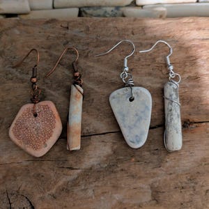 May include: Four pairs of handmade earrings. The earrings feature natural stone and copper or silver wire. One pair has a peach-colored, heart-shaped stone with a textured pattern. The other pairs have rectangular and teardrop-shaped stones.