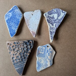 May include: Five ceramic pendants with drilled holes, ideal for jewellery making. The pendants display diverse patterns, including blue speckled, floral, and textured designs. The colour scheme includes blue, white, brown, and black.