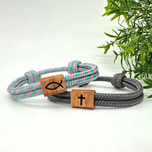 Può includere: Due braccialetti in corda intrecciata con ciondoli in legno. Un braccialetto è grigio e ha un ciondolo in legno a forma di croce. L'altro braccialetto è rosa e verde e ha un ciondolo in legno a forma di pesce.